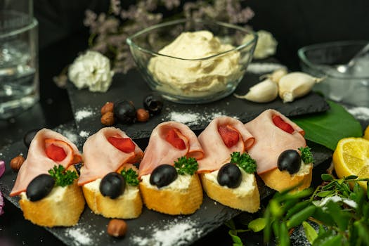 Bouchées apéritives sur pain avec jambon, olives et crème, entourées de fleurs et d'ingrédients frais.